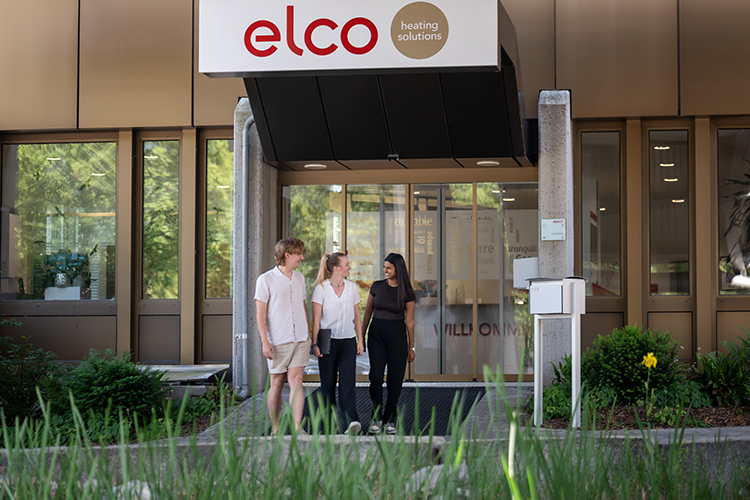 Drei Mitarbeitende vor dem Eingangsbereich eines ELCO Unternehmensstandorts mit sichtbar angebrachtem Firmenlogo.